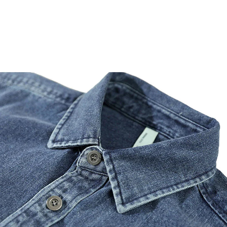 Close-up of a blue washed denim overshirt crafted from natural cotton, featuring a pointed collar and two visible buttons, shown on a white background.