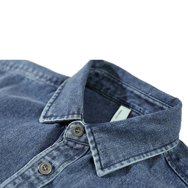 Close-up of a blue washed denim overshirt crafted from natural cotton, featuring a pointed collar and two visible buttons, shown on a white background.