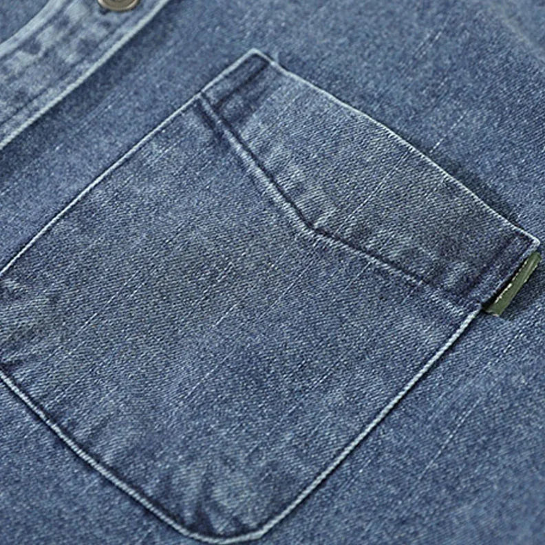 Close-up of a blue washed denim overshirt pocket with visible stitching and a metal button at the top edge, highlighting the natural cotton texture and weave of the fabric.