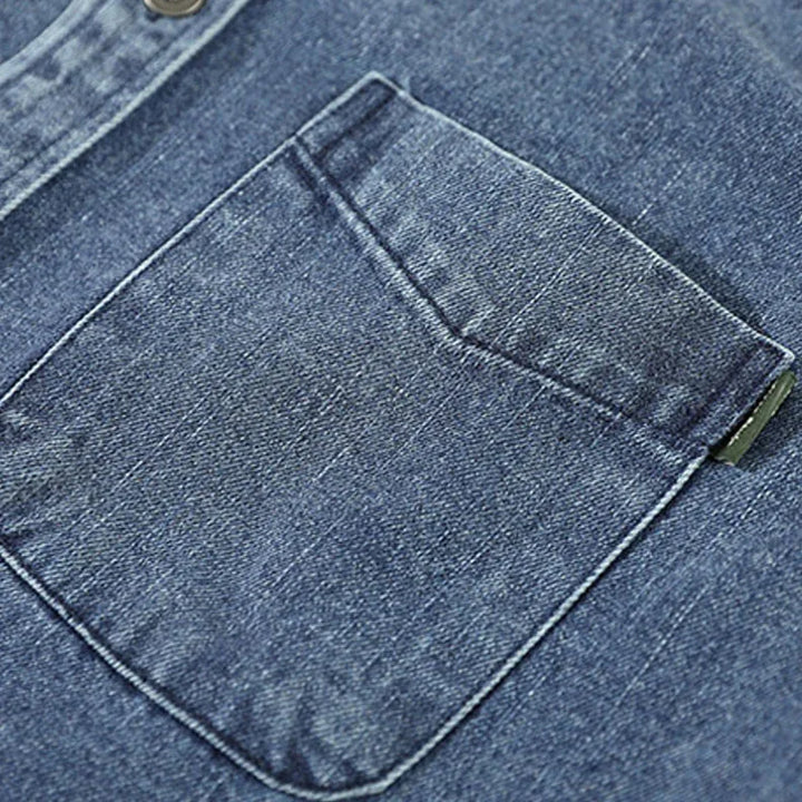 Close-up of a blue washed denim overshirt pocket with visible stitching and a metal button at the top edge, highlighting the natural cotton texture and weave of the fabric.