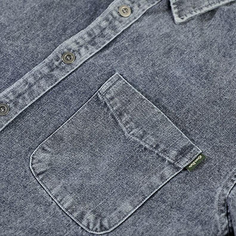 Close-up of a blue washed denim overshirt featuring a chest pocket with a small green tag and visible buttons along the natural cotton shirt placket and collar.