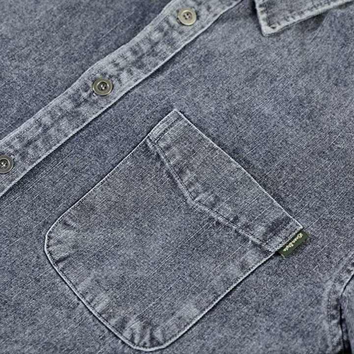 Close-up of a blue washed denim overshirt featuring a chest pocket with a small green tag and visible buttons along the natural cotton shirt placket and collar.