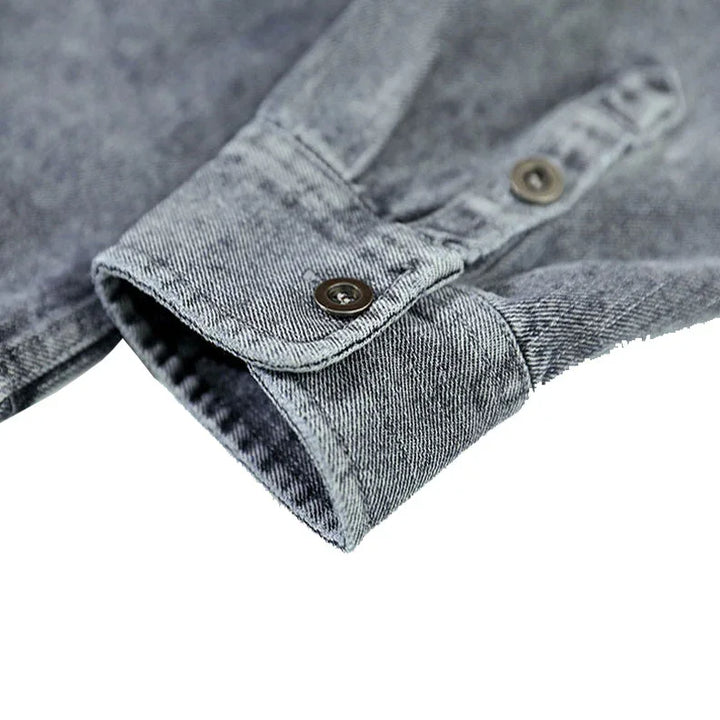 Close-up of a gray washed denim overshirt cuff with two metal buttons, highlighting the texture and stitching details of natural cotton. The background is white.