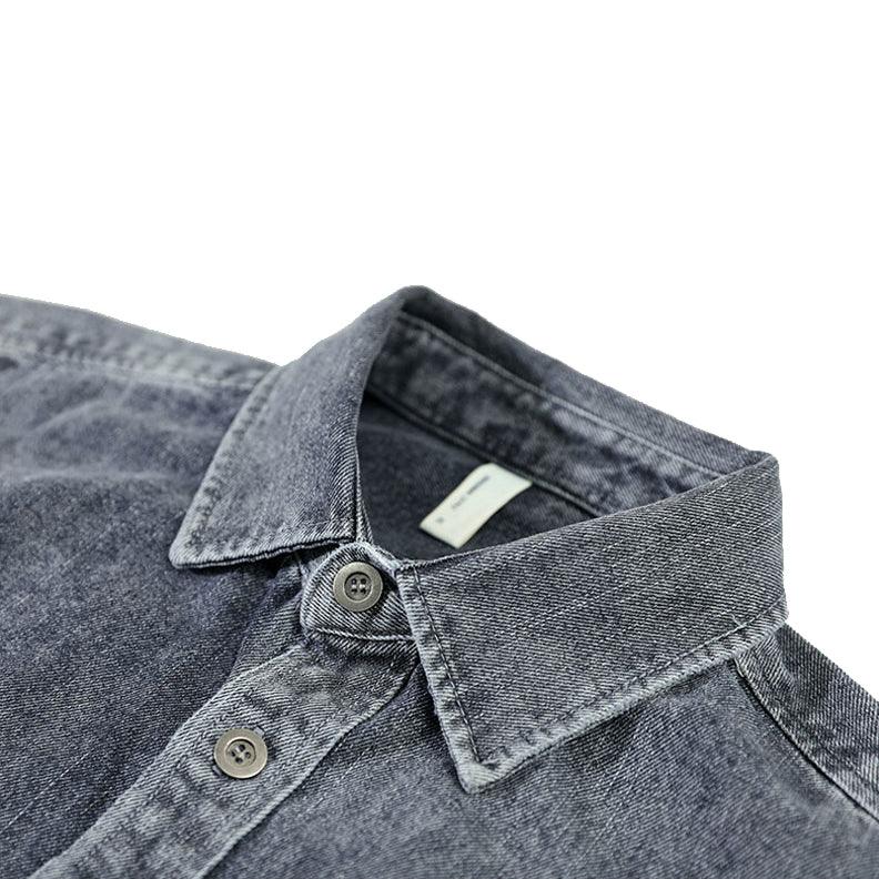 A close-up view of a dark grey washed denim overshirt with a pointed collar and two buttons visible. The shirt has visible stitching, is made from natural cotton, and features a slightly faded, textured appearance.