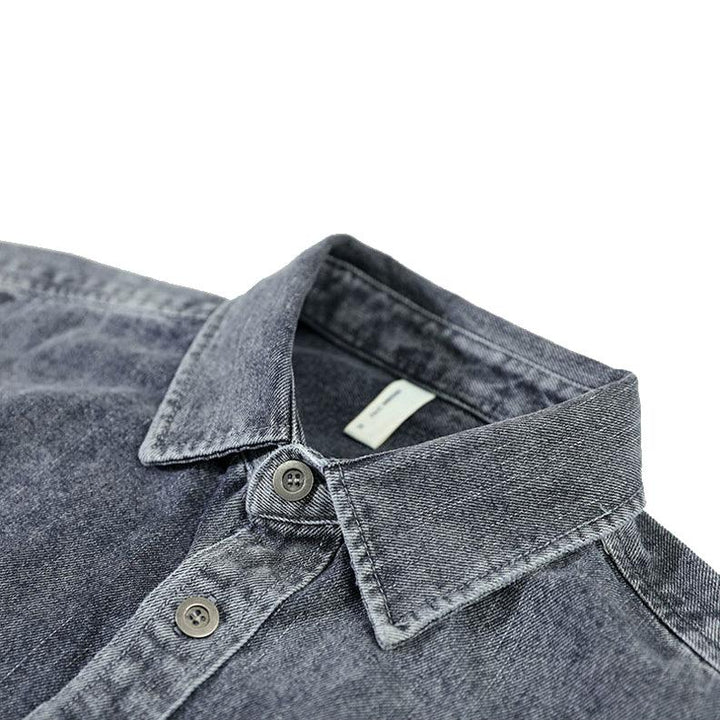 A close-up view of a dark grey washed denim overshirt with a pointed collar and two buttons visible. The shirt has visible stitching, is made from natural cotton, and features a slightly faded, textured appearance.