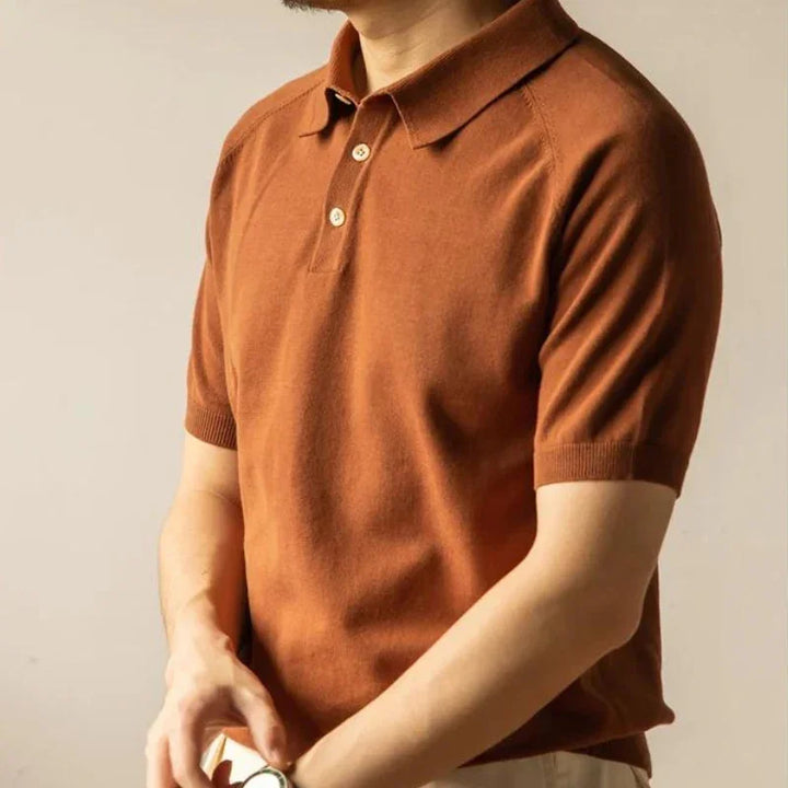A man wearing a classic style polo in a rust color with beige pants stands against a plain background, adjusting something on his wrist. His face is not visible.