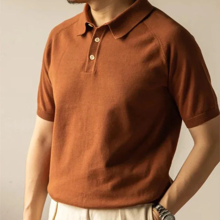 A person wearing a rust-colored organic cotton polo and light beige pants, with one hand in their pocket and a watch visible on their wrist. The persons face is not shown.