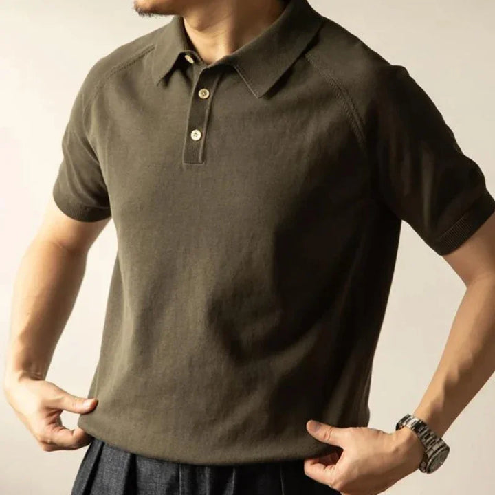 A person wearing a short-sleeved, olive green mens polo shirt and a wristwatch stands with their hands pulling slightly at the sides of the shirt. The face is not visible.