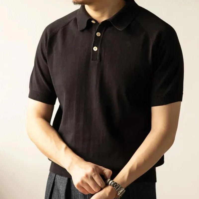 A person wearing a short-sleeve black organic cotton polo with a collar and buttons, standing with arms crossed and hands clasped, dressed in dark pants and a watch. The face is not shown.