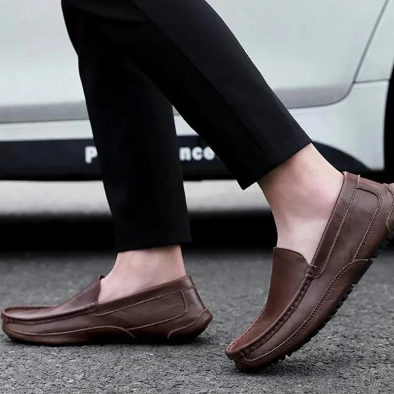 A person wearing black pants and genuine leather loafers with a cushioned insole is walking on a paved street, with a white car visible in the background.