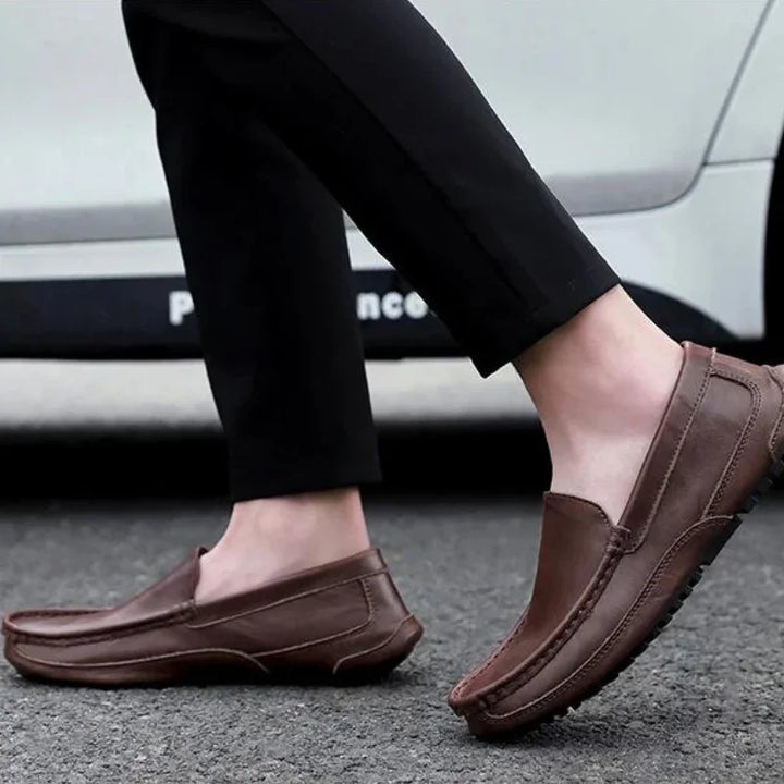 A person wearing black pants and genuine leather loafers with a cushioned insole is walking on a paved street, with a white car visible in the background.