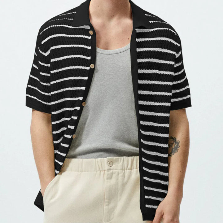 A person wearing a black short-sleeve button-up shirt with white horizontal stripes, styled streetwear over a light grey undershirt and beige pants. Their hands are in their pockets, with part of a tattoo visible on their left arm.
