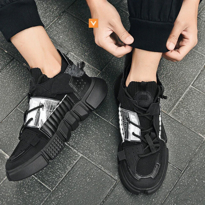 A person wearing black and white Extreme VS7 Sneakers with thick soles, adjusting the cuff of their black pants while standing on a gray tiled floor.