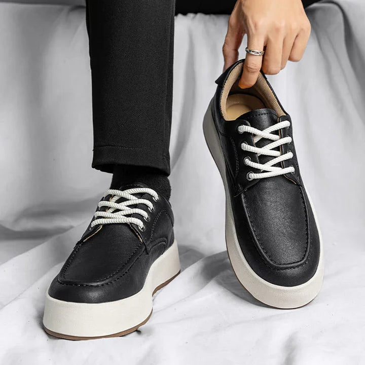 A person wearing black pants and socks is putting on a pair of Men's Leather Casual Shoes with white laces and thick white soles, seated on a light-colored fabric background.