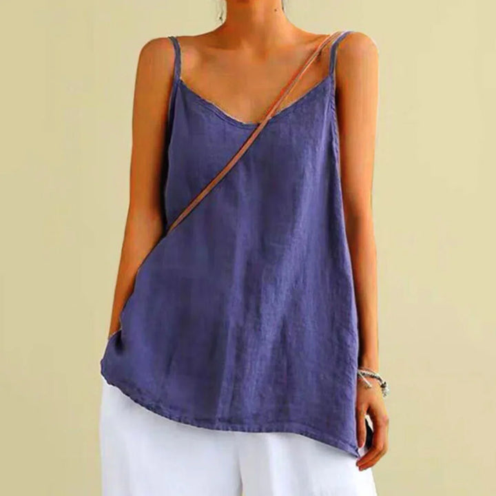 A person wearing a loose, sleeveless urban-chic camisole with thin straps and white pants stands against a beige background. A thin brown strap crosses diagonally over the shoulder and chest.