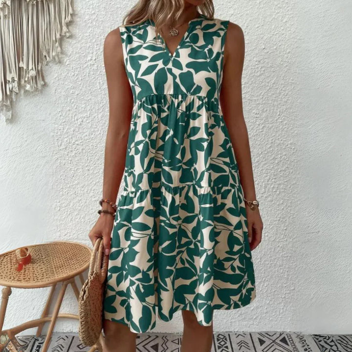 A woman stands against a white textured wall, wearing a sleeveless dress in a lightweight breathable fabric with a green leaf pattern—an ideal addition to your summer wardrobe. She stands by a wicker chair with a woven bag and macramé wall hanging nearby.