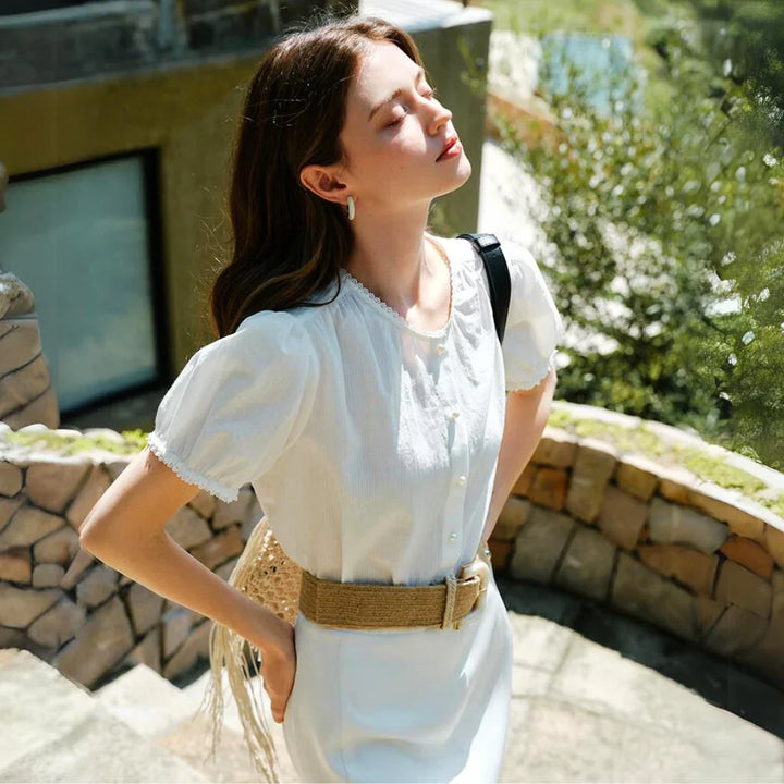 A woman in a white lace trim blouse and dress stands on stone steps outdoors, eyes closed and head tilted back, enjoying the sunlight. She wears a wide beige belt, gold earrings, and carries a bag over her shoulder. Greenery surrounds the scene.