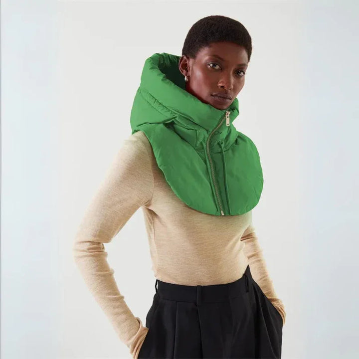 A person wearing a beige long-sleeve top, black trousers, and a lightweight puffy green hooded scarf with a zip-up front, standing against a plain white background.