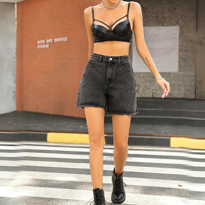 A person crossing the street in a black leather bralette, high-waist black wash denim streetwear shorts, black ankle boots, and a necklace, with a brick building and crosswalk visible in the background.