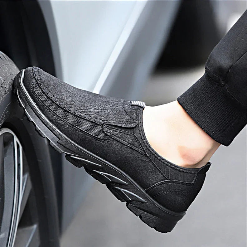 A person wearing breathable loafers with textured uppers and thick soles rests their foot against a car tire. The comfortable fit complements their black pants, perfect for a stylish, laid-back look.