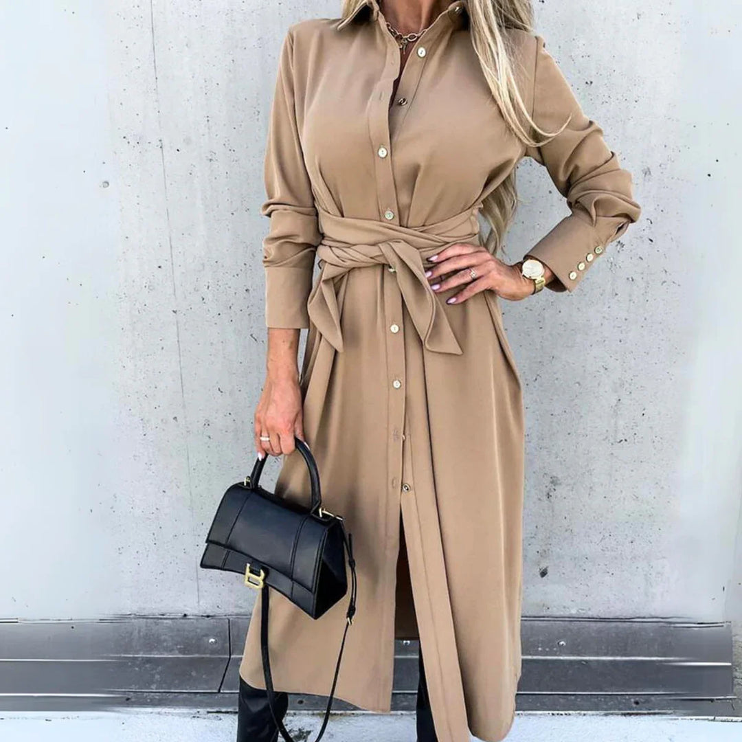 A woman wearing a long-sleeve wrap dress in tan with a waist tie, holding a black handbag and wearing a watch, stands against a concrete wall. Her long blonde hair is styled loosely, capturing effortless high fashion streetwear vibes.