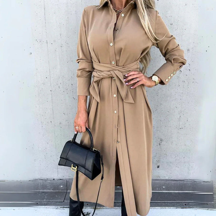 A woman wearing a long-sleeve wrap dress in tan with a waist tie, holding a black handbag and wearing a watch, stands against a concrete wall. Her long blonde hair is styled loosely, capturing effortless high fashion streetwear vibes.
