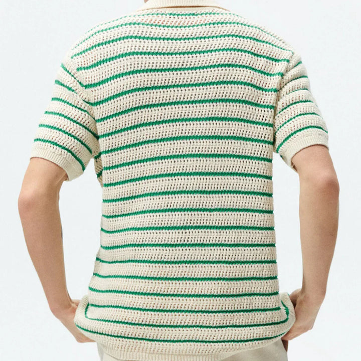 A person is shown from behind, wearing a short-sleeved, crocheted shirt with an open-knit design and horizontal green and cream stripes. Their hands rest on their hips. The background is plain and light-colored.