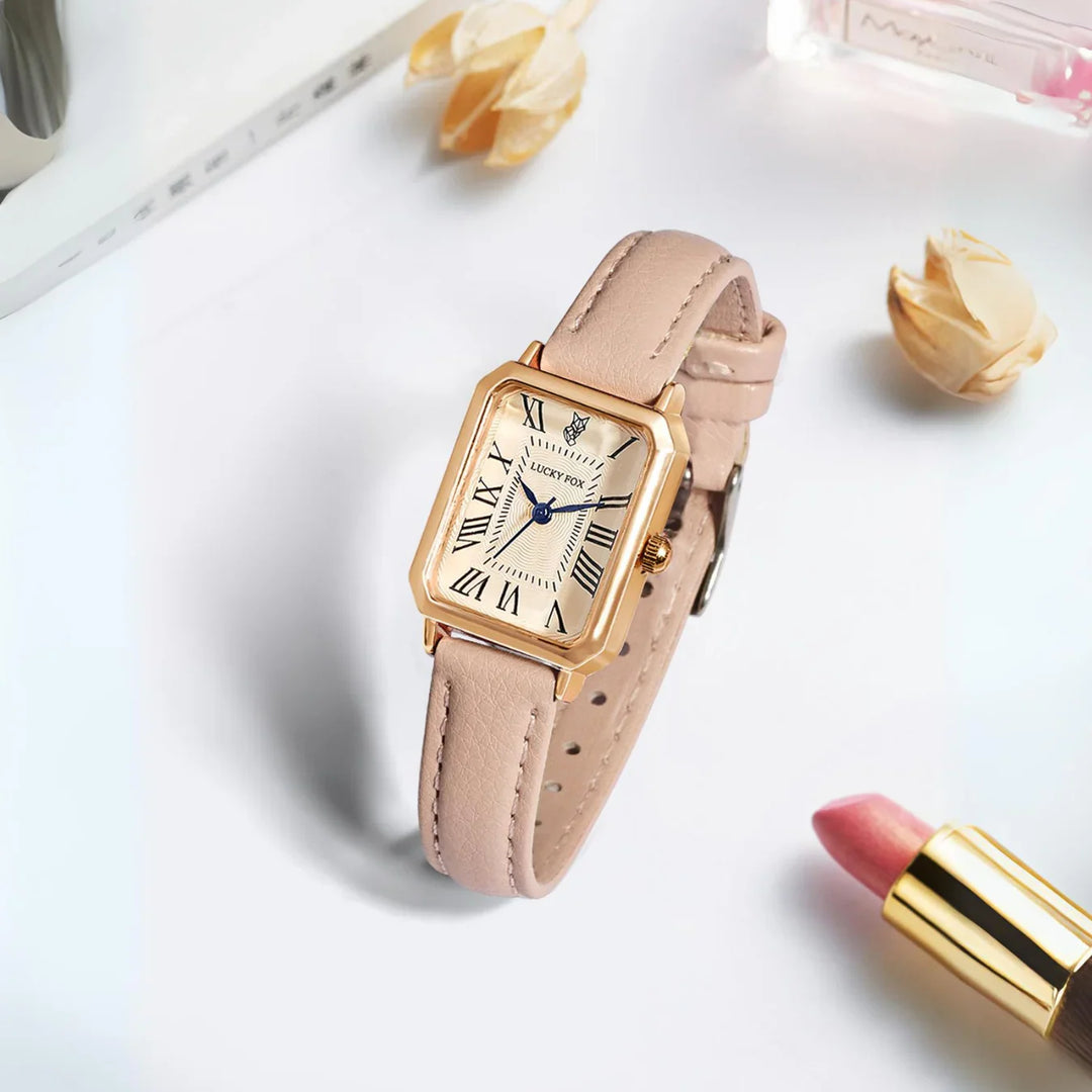 A gold rectangular face watch with a genuine leather strap rests on a white surface, surrounded by pink lipstick, perfume, and dried flowers. The watch features elegant Roman numerals and striking blue hands.