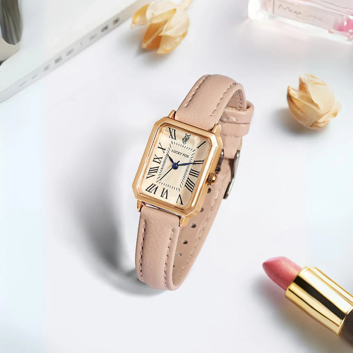 A gold rectangular face watch with a genuine leather strap rests on a white surface, surrounded by pink lipstick, perfume, and dried flowers. The watch features elegant Roman numerals and striking blue hands.