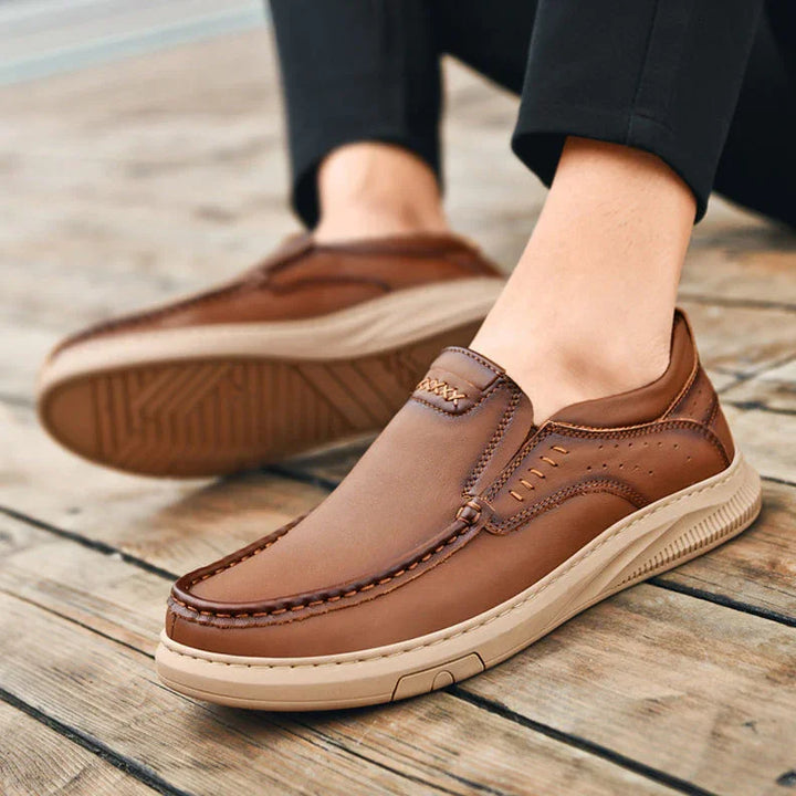 A person wearing men's cowhide leather shoes with soft soles sits on a wooden surface. These slip-on loafers feature detailed stitching and offer a casual, comfortable design.