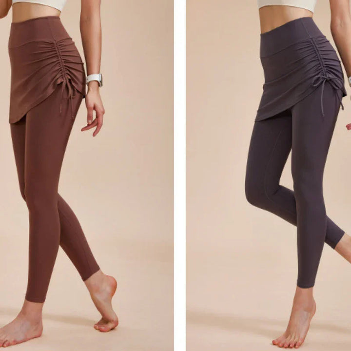 Two women model high-waisted leggings with skirt, featuring ruched detailing in brown and dark gray—an ideal addition to your autumn wardrobe. They stand barefoot against a neutral beige background.