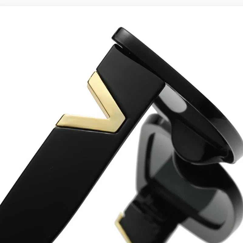 Close-up of the black temple arm of designer sunglasses, featuring a gold V-shaped accent. The lens and part of the geometric frame are partially visible in the background.