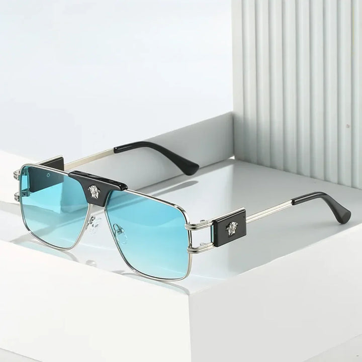 A pair of bold sunglasses with rectangular, blue-tinted lenses and premium metal frames, featuring black accents and a metallic emblem on the temples, displayed on a white geometric surface.