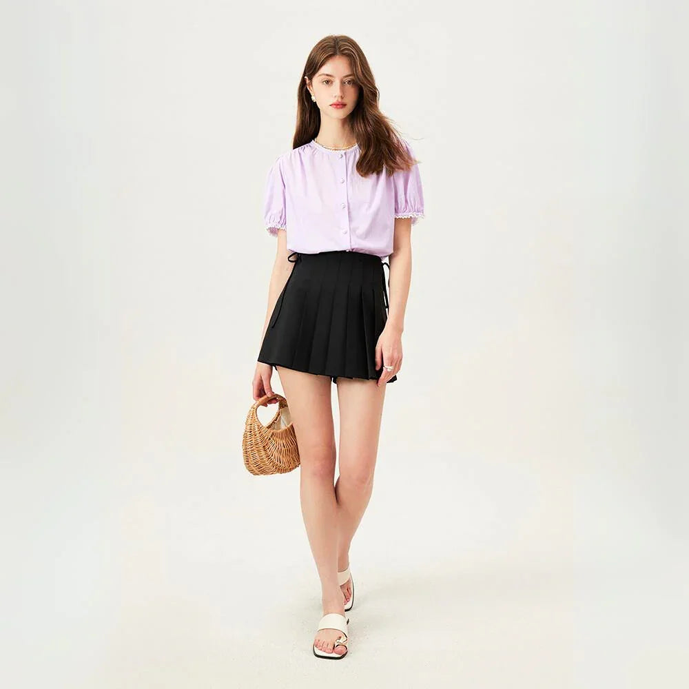 A woman stands against a plain light background, wearing a versatile blouse in light purple, a black pleated mini skirt, white sandals, and holding a small woven handbag. She has long brown hair and a relaxed expression.