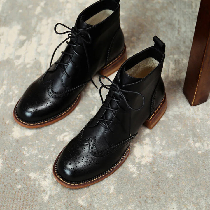 A pair of black leather brogue boots with decorative perforations and brown soles, crafted from genuine leather, placed on a light textured surface next to a wooden furniture leg.