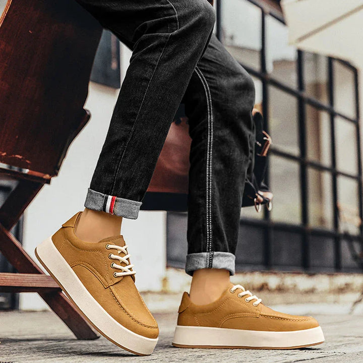 A person wearing cuffed black jeans and tan men's leather casual shoes with white soles sits on a wooden bench outdoors near large windows.