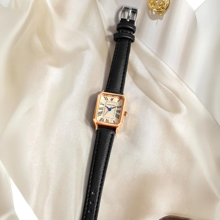 A rectangular face watch with a genuine leather strap and Roman numerals, displayed on smooth, cream-colored satin fabric near a gold decorative accessory.