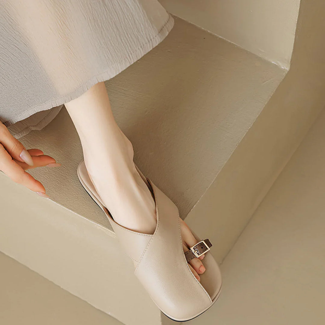 A person wearing a light gray dress is seated on a beige step, showing their feet in chic slides—beige open-toe sandals with a buckle detail—perfect for elevating any summer wardrobe.