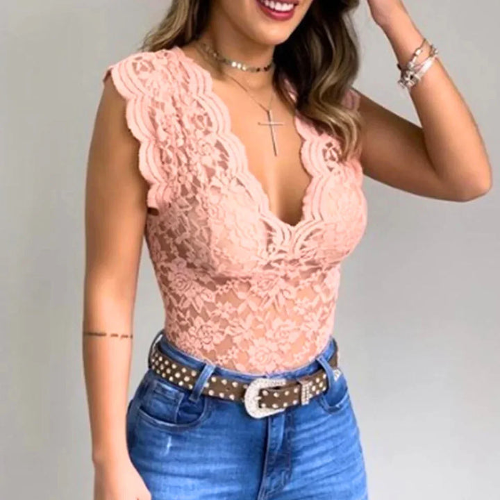 A woman wearing a sleeveless peach-colored lace top with a deep V-neck and scalloped edges, blue jeans with a studded belt, layered necklaces, and bracelets. She is smiling and touching her hair, showcasing effortless summer fashion.