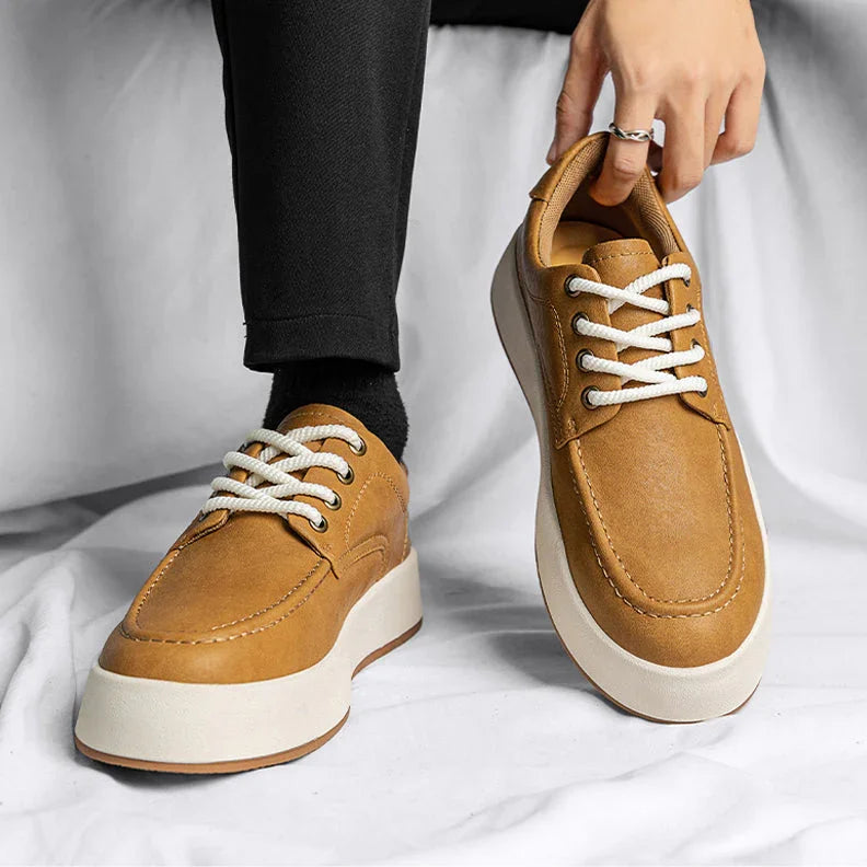 A person wearing black pants and socks sits on a white surface, holding one tan lace-up sneaker with white soles—stylish men's leather casual shoes—while the other sneaker is worn on their left foot.