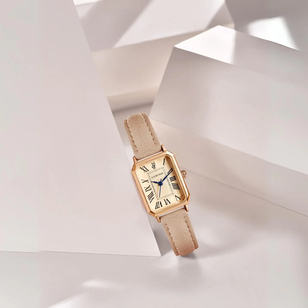 A rectangular face watch with a beige genuine leather strap, Roman numeral markers, and blue hands is displayed among abstract white geometric shapes with soft lighting and shadows.