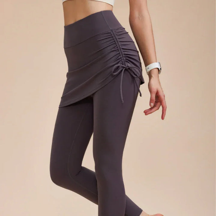 A woman wearing high-waisted leggings with skirt in dark purple and a white smartwatch, standing against a beige background—an ideal pick for your autumn wardrobe.