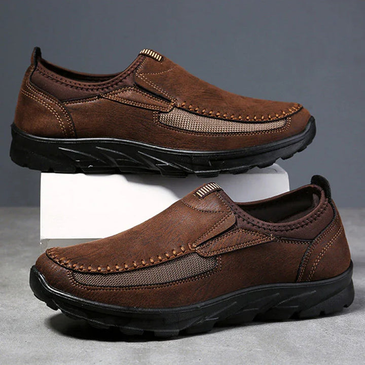 A pair of brown Men Leather Casual Shoes with black rubber soles and visible stitching, designed for a comfortable fit. Displayed against a gray background, one breathable loafer is propped up on a white block above the other.