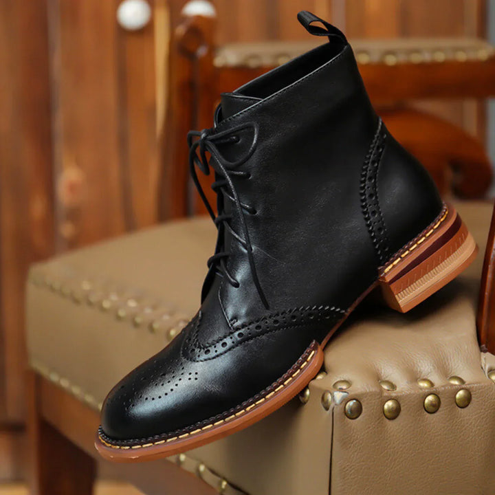 A black leather brogue boot with lace-up design and brown sole is displayed on a tan, gold-studded upholstered chair in a warmly lit room—perfect for the urban trendsetter seeking genuine leather style.