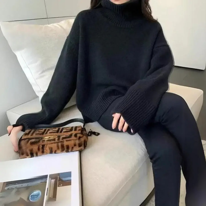 A person sits on a white couch, wearing a loose black cashmere turtleneck sweater and black pants. Their hand rests on a brown patterned pouch, and a magazine lies on the table in front of them.