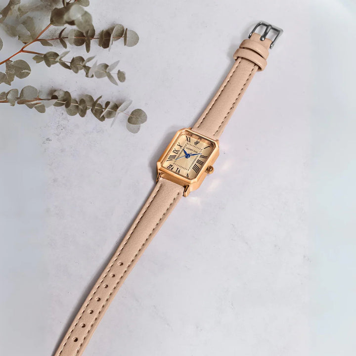 A gold Roman numeral watch with a rectangular face and a genuine leather strap lies on a light marble surface, next to a sprig of dried eucalyptus leaves in the top left corner.