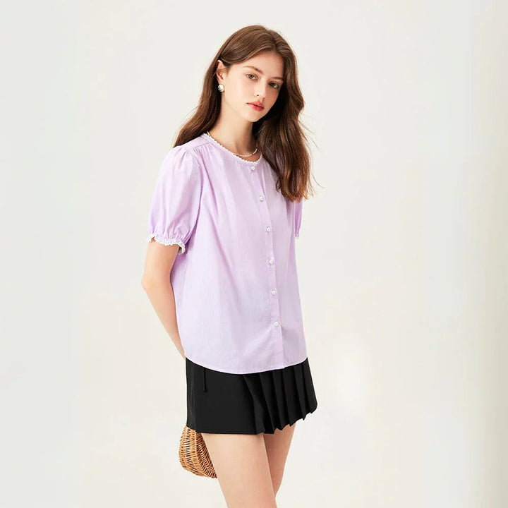 A woman with long brown hair wears a light purple versatile blouse with short puff sleeves and a black pleated skirt, standing against a plain light background, holding a small woven bag behind her back.
