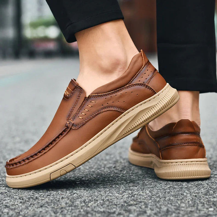 Person wearing men's cowhide leather slip-on shoes with beige soft soles, standing on a paved outdoor surface. The shoes feature decorative stitching and a casual design. Only the lower legs and feet are visible.