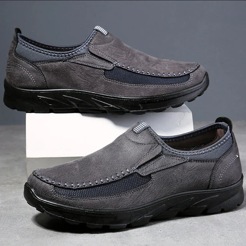 A pair of gray Men Leather Casual Shoes with black soles and stitching details are displayed, one shoe resting on a white block and the other flat on a gray surface, offering a comfortable fit against a plain gray background.