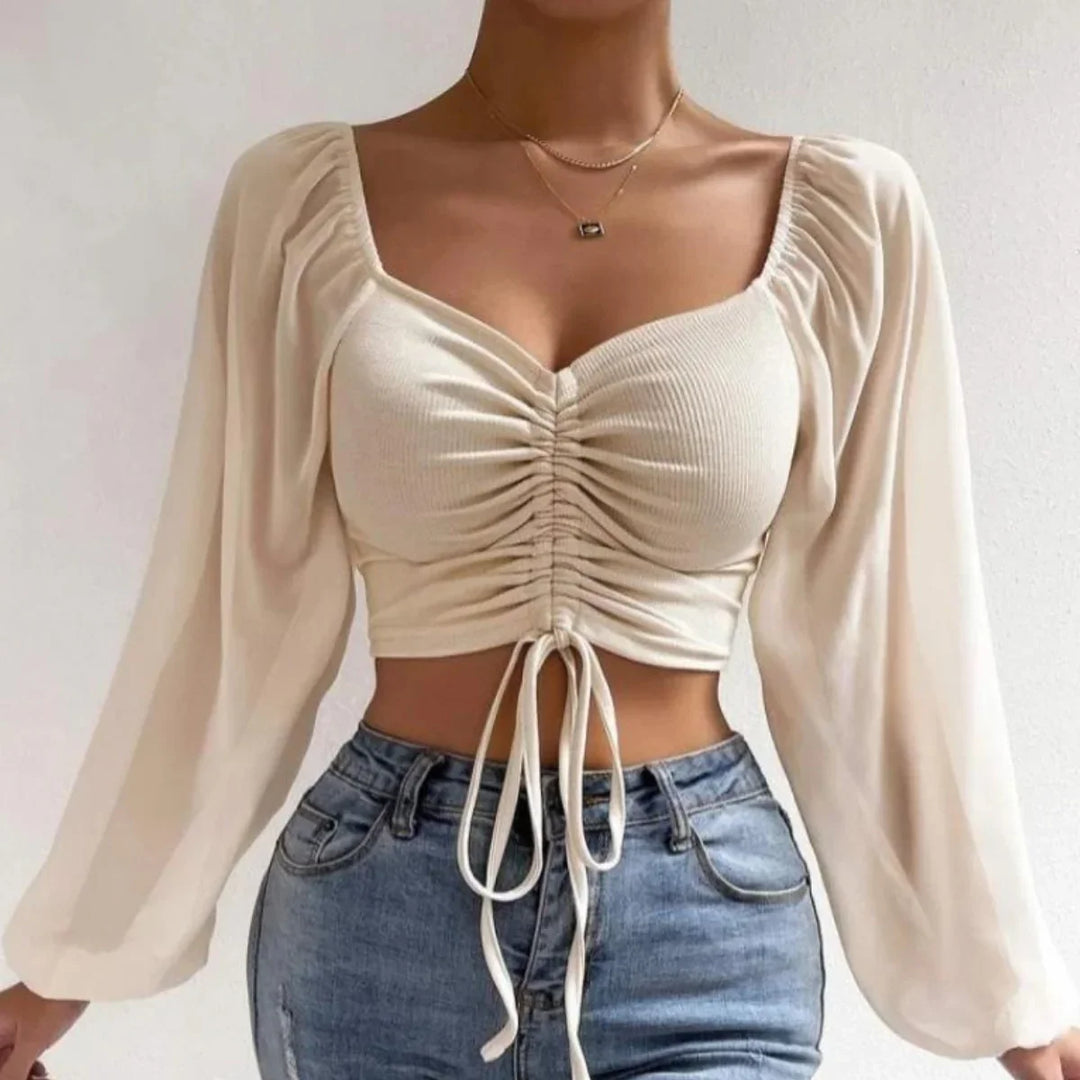 A woman wearing a cream-colored, long-sleeve crop top with a ruched front and drawstring, blue high-waisted jeans, and layered necklaces stands against a plain light background.